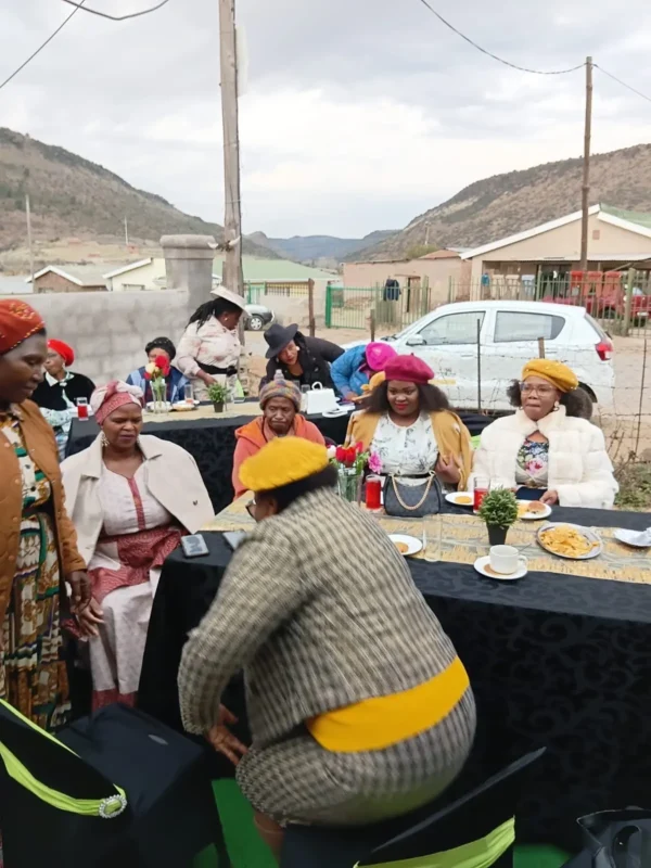 Women seated at decorated tables in a walled yard in Burgersdorp, with the Stormberg hills behind.